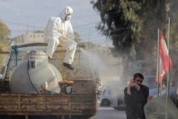 A municipal worker wearing protective gear sprays disinfectant as a precaution against the coronavirus outbreak as a man speaks on his mobile phone, in the suburb of Beir Hassan, Beirut, Lebanon, Monday, March 16, 2020. (AP Photo/Hassan Ammar)