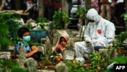 Seorang anggota keluarga mengenakan pakaian pelindung diri (APD) bersama dua anak berdoa di dekat makam korban COVID-19 yang baru saja dimakamkan, di Lhokseumawe, Aceh, 18 Mei 2021. (Azwar Ipank / AFP)