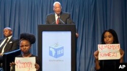 Protesters hold up signs as President Jacob Zuma speaks at the announcement of the results of municipal elections in Pretoria, South Africa, Aug. 6, 2016. The protest refers to Zuma's acquittal on rape charges in 2006.
