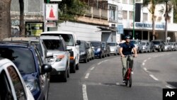 FILE - A cyclist pedals through an empty street, past motorists waiting to fill up at one of the few fuel stations that has electricity, during rolling blackouts in Caracas, Venezuela, March 10, 2019. On Friday, Venezuelans waited hours in line for gasoline.