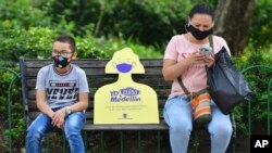 In this June 8, 2020 photo, a sign reminding citizens to maintain a safe social distance sits on a park bench between a woman and child, amid the new coronavirus pandemic in Medellin, Colombia.