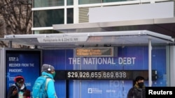 People wearing protective face masks wait at a bus stop with a display of the current national debt amid the COVID-19 pandemic in Washington, Jan. 31, 2022. 