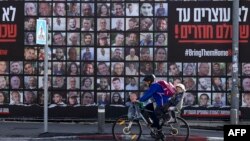 Seorang pria bersepeda bersama seorang anak melewati papan reklame bergambar warga Israel yang disandera di Gaza sejak serangan Hamas pada 7 Oktober 2023, di Yerusalem, 15 Januari 2025. (Foto: AFP)