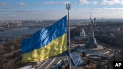 Bendera nasional Ukraina berkibar di dekat Monumen Tanah Air (kanan), di Kyiv, Minggu, 13 Februari 2022. (AP/Efrem Lukatsky)