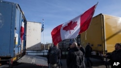 Demonstrators, one holding a Canadian flag, stand next some of the 20 trucks which blocked one entrance to the government buildings, rear, in The Hague, Netherlands, Saturday, Feb. 12, 2022, to protest against COVID-19 restrictions.