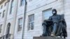 FILE - The John Harvard statue at Harvard University, a popular tourist attraction at the campus in Cambridge, Masssachusetts, sits adorned with a mask, March 14, 2020. The university is restricting spending in the wake of the coronavirus outbreak.