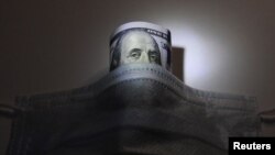 Selembar uang kertas dollar AS di balik masker untuk melindungi dari virus corona (Covid-19), dalam foto ilustrasi pada 18 Maret 2020. (Foto: Reuters)