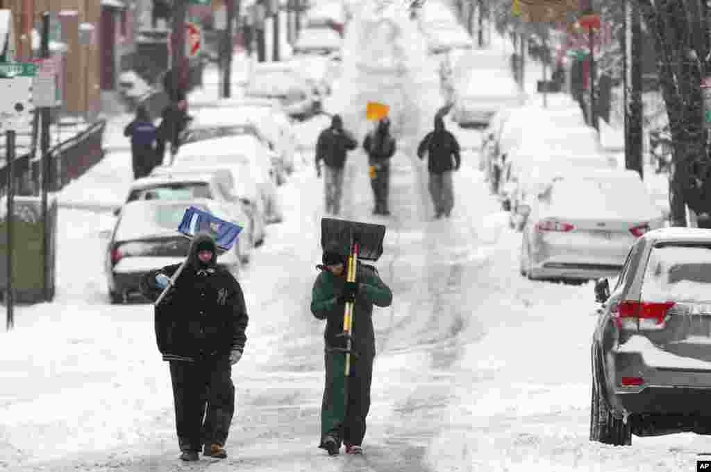 Para pria berjalan membawa pengki di tengah jalanan yang ditutupi salju setelah badai salju di Hoboken, New Jersey (27/1). ​(AP/Julio Cortez)