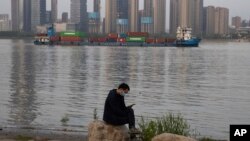 In this Monday, April 13, 2020, photo, a man wearing a mask against the new coronavirus checks his phone as a container ship cruises along the Yangtze River in Wuhan in Central China's Hubei province. China's exports fell further in March compared…