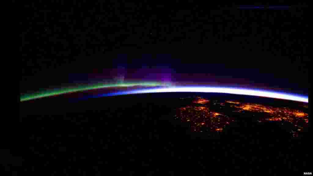 Night lights from the International Space Station show cities of Ireland and the United Kingdom in the foreground contrasted by the bright sunrise in the background. The greens and purples of the Aurora Borealis are seen along the rest of the horizon. 