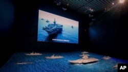 A TV screen showing the U.S. Navy fleet sail in formation near the models of Liaoning aircraft carrier with navy frigates and submarines on display at the military museum in Beijing, Thursday, Aug. 1, 2019. U.S. Secretary of State Mike Pompeo said…