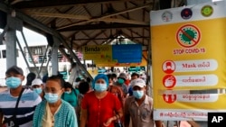 Penumpang feri mengenakan masker di tengah pandemi COVID-19, berjalan melewati poster "Hentikan COVID-19" di dermaga Pansodan, Yangon, Myanmar. (AP)