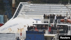 Para penyintas dan keluarga korban musibah kapal Costa Concordia berdiri di dalam sebuah kapal ferry yang berhenti di dekat kapal pesiar yang teronggok di dekat pelabuhan Giglio, Italia, untuk memperingati setahun tenggelamnya kapal pesiar tersebut (13/1).